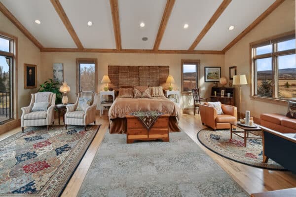 Spacious bedroom with vaulted wooden beam ceiling, large bed centered against a woven headboard, armchairs, leather sofas, rugs, and large windows offering scenic views. Warm, cozy decor perfect for a ranch for sale or hunting property.