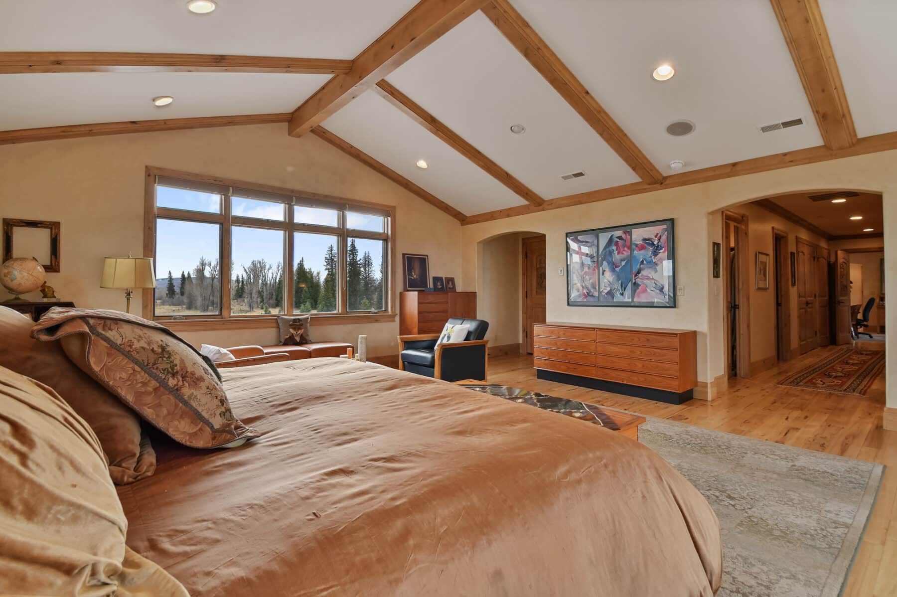Spacious bedroom with wooden beams on the ceiling, large window with outdoor view, tan bedding, art on the walls, dressers, armchair, and light hardwood floors—perfect for a cozy retreat on a ranch for sale or recreational land.