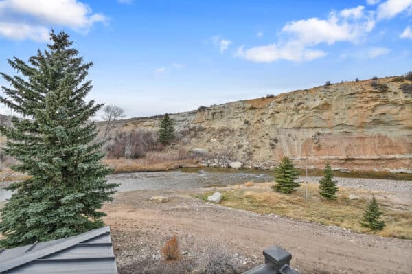 A scenic view of a rocky hillside beside a shallow river, with evergreen trees scattered along the riverbank and a partly cloudy blue sky overhead—ideal land for sale for those seeking peaceful hunting property.
