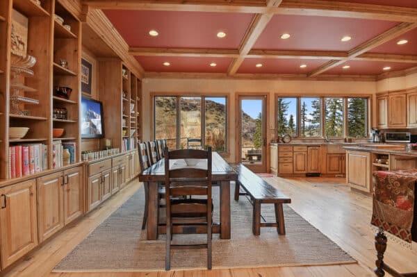 A rustic kitchen and dining area with wooden cabinets, a large table and bench, shelves with books and dishes, and large windows showing mountain views—perfect for enjoying ranch for sale or recreational land living. Recessed lighting adds to the cozy interior.