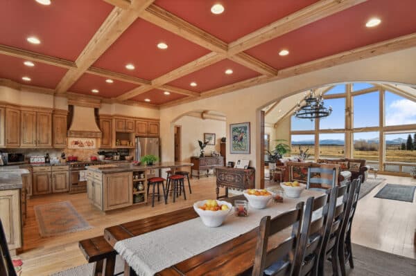 Spacious open-concept kitchen and dining area in this ranch for sale features wood cabinets, island, rustic dining table, and large windows offering scenic views of a natural landscape under a reddish coffered ceiling with recessed lighting.