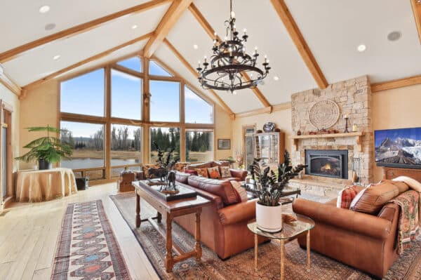 Spacious living room with large windows, leather sofas, a stone fireplace, wooden ceiling beams, indoor plants, a chandelier, and scenic views of trees and water—perfect for enjoying your ranch for sale or recreational land.