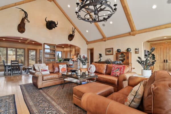 Spacious living room with brown leather sofas, patterned rug, vaulted ceiling, and view into dining area and kitchen. Wooden accents and large windows enhance the warm, rustic atmosphere of this inviting cattle ranch.
