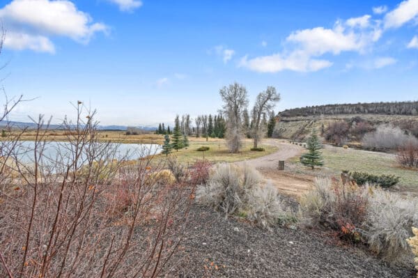 A dirt path winds through a landscape with sparse trees and shrubs, alongside a pond under a bright blue sky. Distant mountains and grassy fields complete this scenic hunting property or ranch for sale.