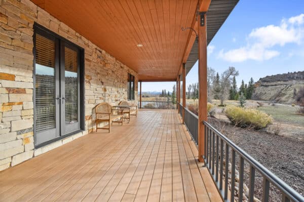 A spacious wooden porch with two chairs overlooks recreational land featuring trees, shrubs, and distant hills under a partly cloudy sky. The house exterior showcases stone walls and glass double doors, ideal for those seeking land for sale.
