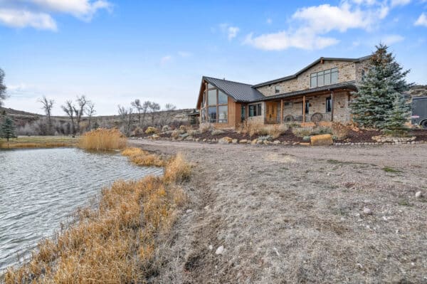 A modern rustic house with stone and wood features sits beside a small pond on recreational land, surrounded by dry grass, rocks, and sparse trees under a partly cloudy sky.
