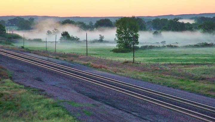 Railroad tracks run alongside a grassy field with scattered trees, as low-lying morning fog drifts near the ground—ideal land for sale. Hills are visible in the distance under a soft, pastel-colored sunrise sky.