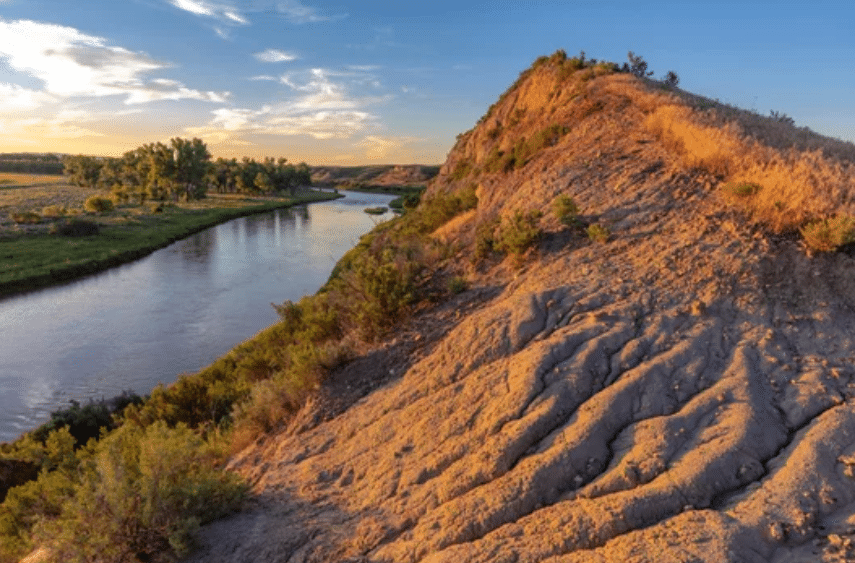 A river winds through a grassy landscape at sunset, beside an eroded, sunlit hill with sparse vegetation—ideal land for sale, perfect for a cattle ranch or recreational land. The sky glows with warm golden light and scattered clouds.