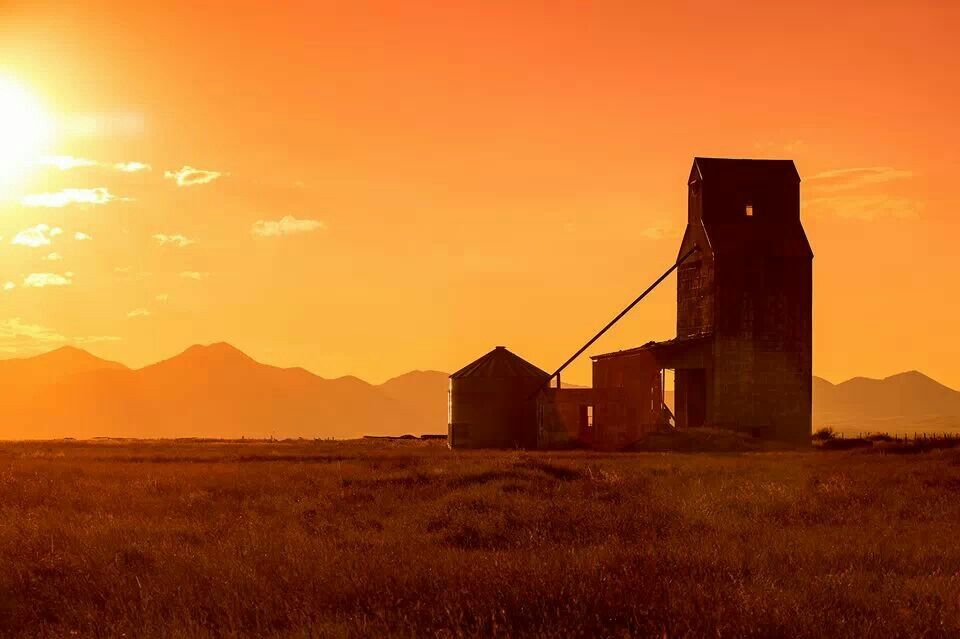 An old, weathered grain elevator stands in a grassy field at sunset, silhouetted against an orange sky with distant mountains—a picturesque view of recreational land perfect for those seeking unique ranch or land for sale.