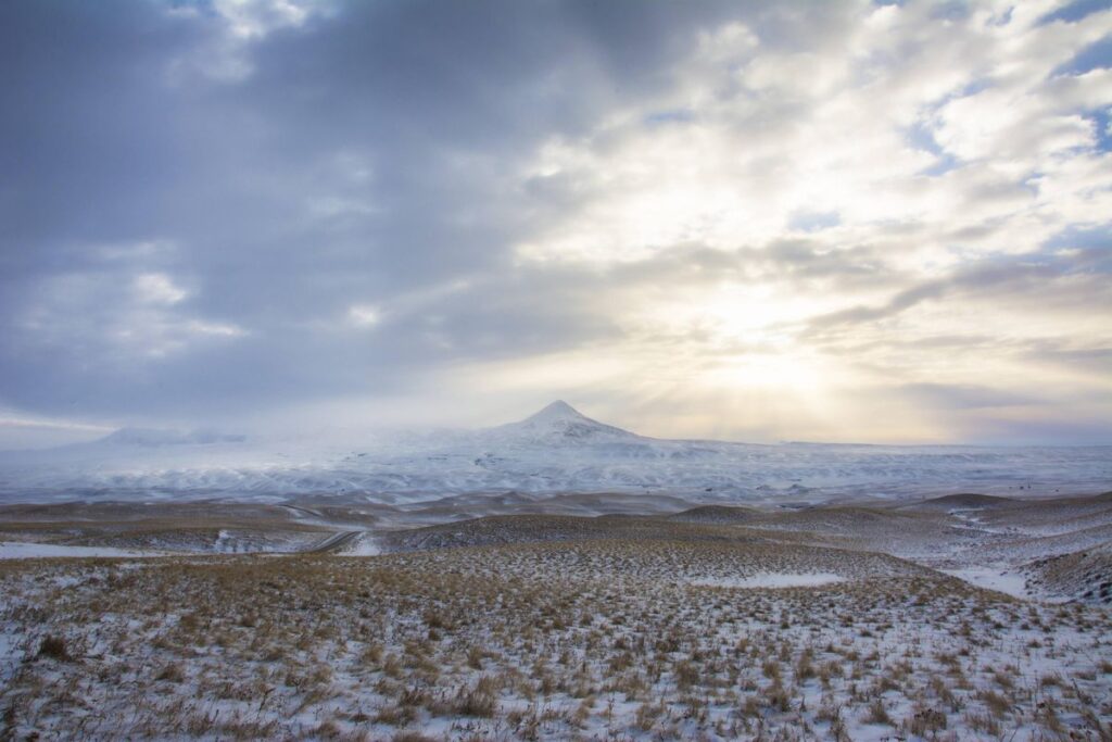Snow-covered plains stretch toward a distant mountain under a cloudy sky, with sunlight illuminating this scenic recreational land. Sparse grasses poke through the snow, highlighting the natural beauty of ranch land for sale.