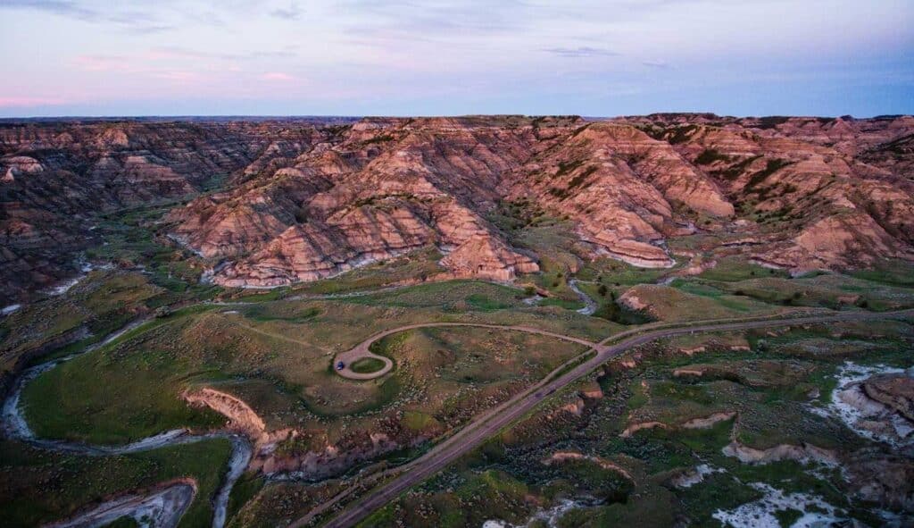 Aerial view of rugged, striped badlands with steep cliffs and green valleys at dusk, intersected by winding roads under a soft pastel sky—ideal land for sale perfect for a cattle ranch.