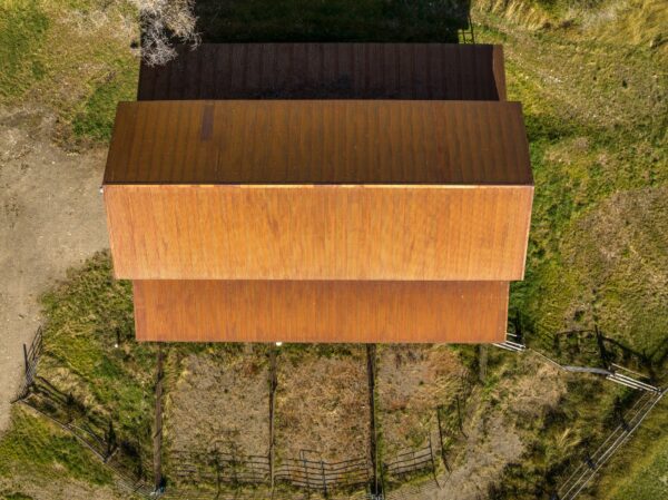 Aerial view of a large rusty-roofed barn surrounded by grass and dirt, with partial fencing and shadows—ideal ranch for sale or hunting property.