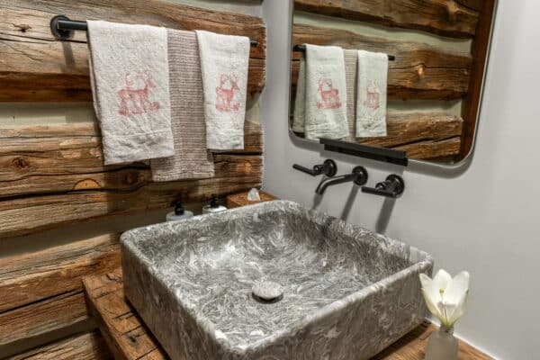 A rustic bathroom with a gray marbled square sink, black wall-mounted faucet, wooden countertop, two hand towels with red animal designs, soap dispensers, mirror, and log cabin-style wooden walls—perfect for a cattle ranch or recreational land retreat.