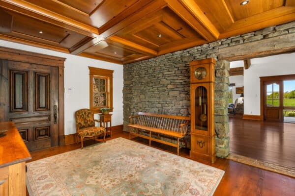 A spacious, rustic entryway sets the tone for this cattle ranch, featuring a wood-paneled ceiling, stone accent wall, grandfather clock, antique bench, armchair, ornate rug, and a large wooden front door. Sunlight streams in from an adjacent room.