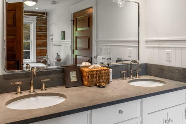 A bathroom with a double sink vanity, gray countertop, and white cabinets—perfect for relaxing after a day on your hunting property. A large mirror reflects the space, with two framed photos, a clock, and a wicker basket on the counter. A bathtub is visible in the background.