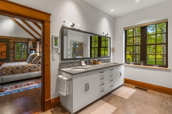 Bright bathroom with a double-sink vanity, large mirror, and two rugs. Window overlooks green trees; door opens to a cozy bedroom with patterned bedding and wood accents in this inviting ranch for sale. Natural light fills both rooms.